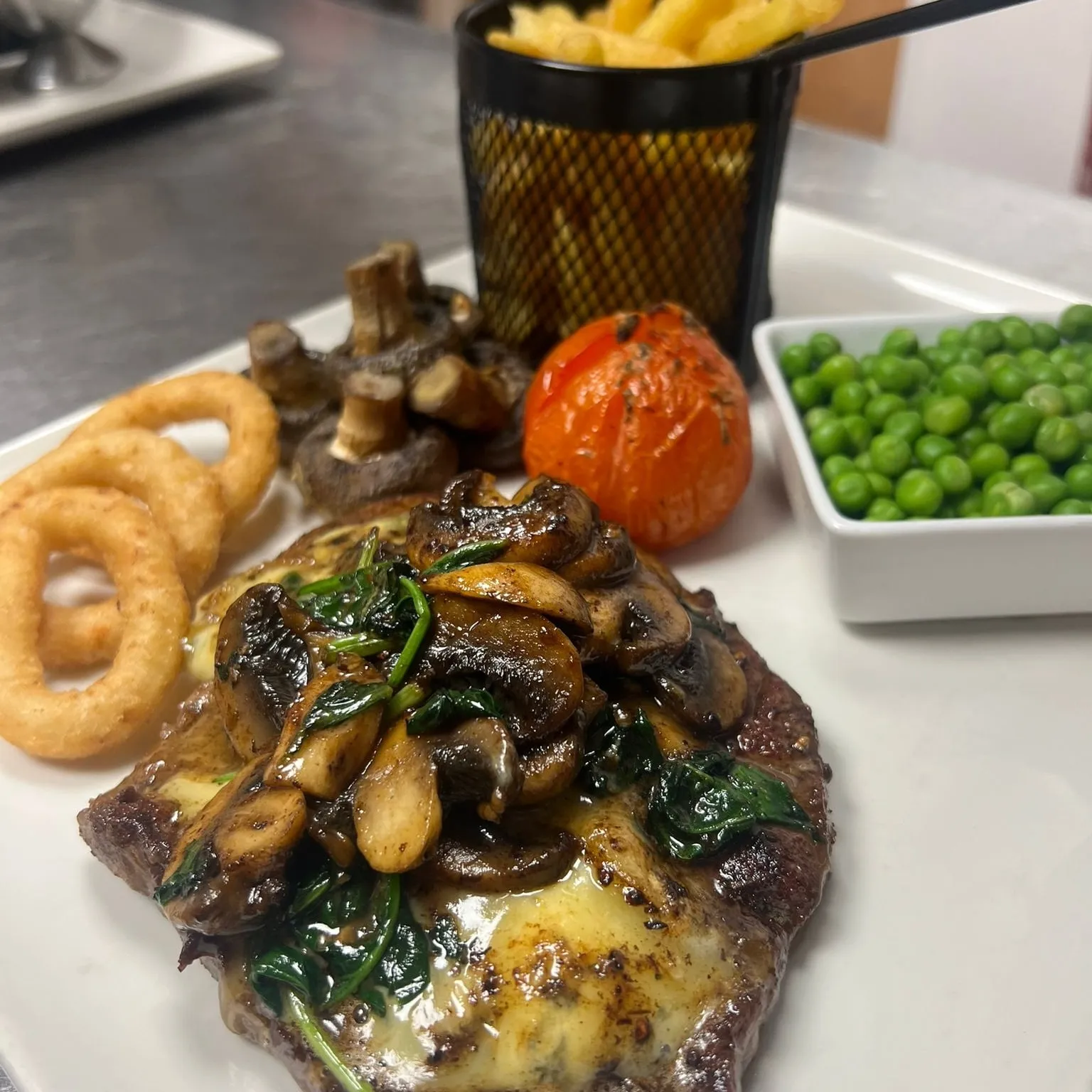 A steak covered in mushrooms with onion rings, chips and peas.